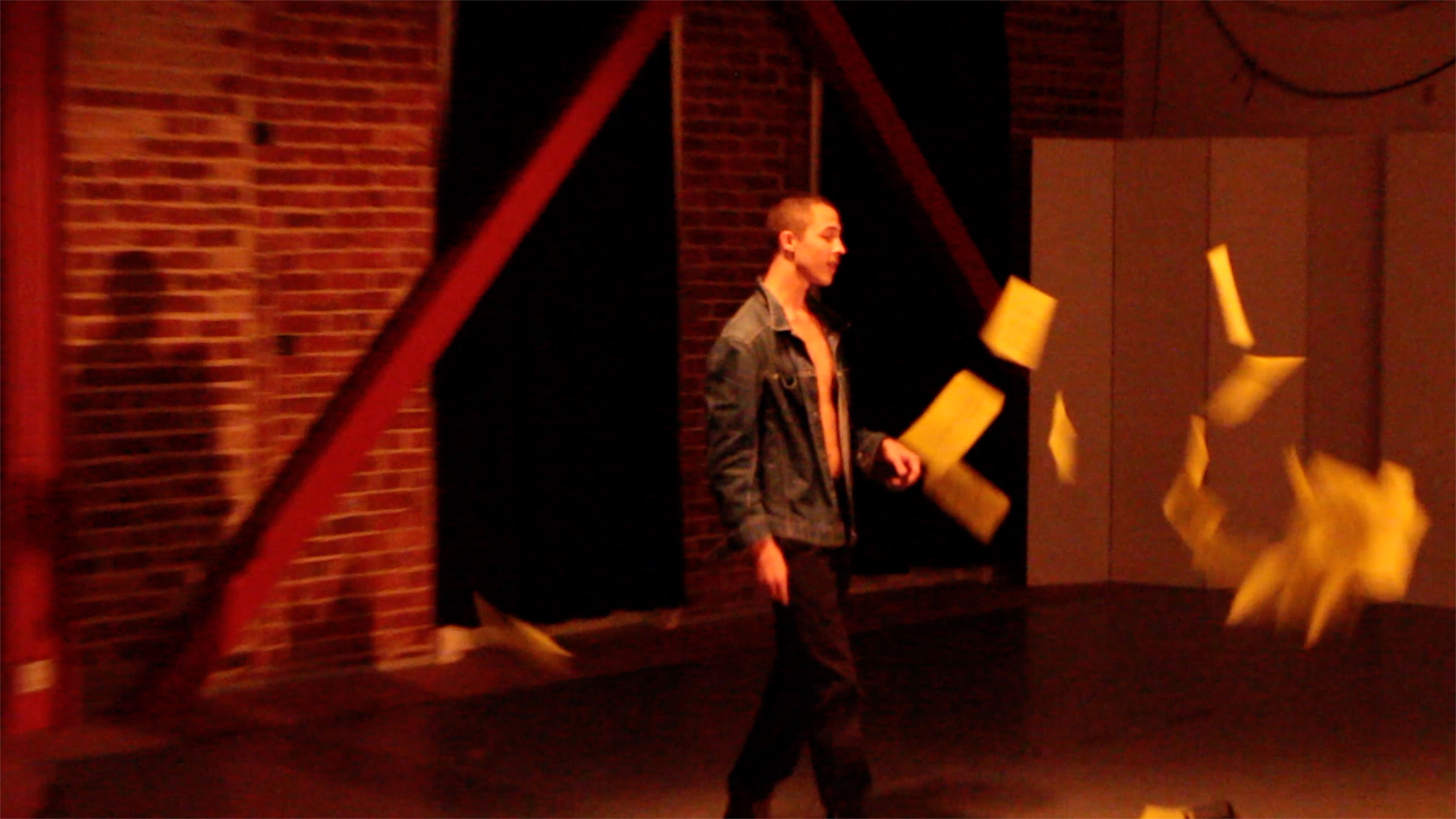 Dalton Alexander stands in a jean jacket, black dickie pants, and boots as yellow papers fly in front of him on their way to the ground.